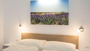 Un dormitorio con una cama, un cuadro colgado en la pared de un campo de flores moradas y dos lámparas de pared.