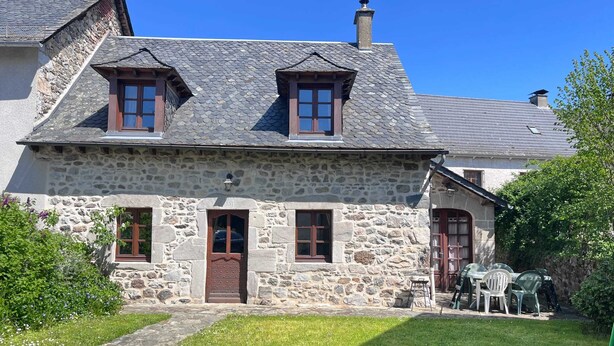 A stone house with a sloped roof, two windows, a door, and a small porch with plastic chairs. - Homerez - Charming House in Lanobre with Garden (Lanobre)