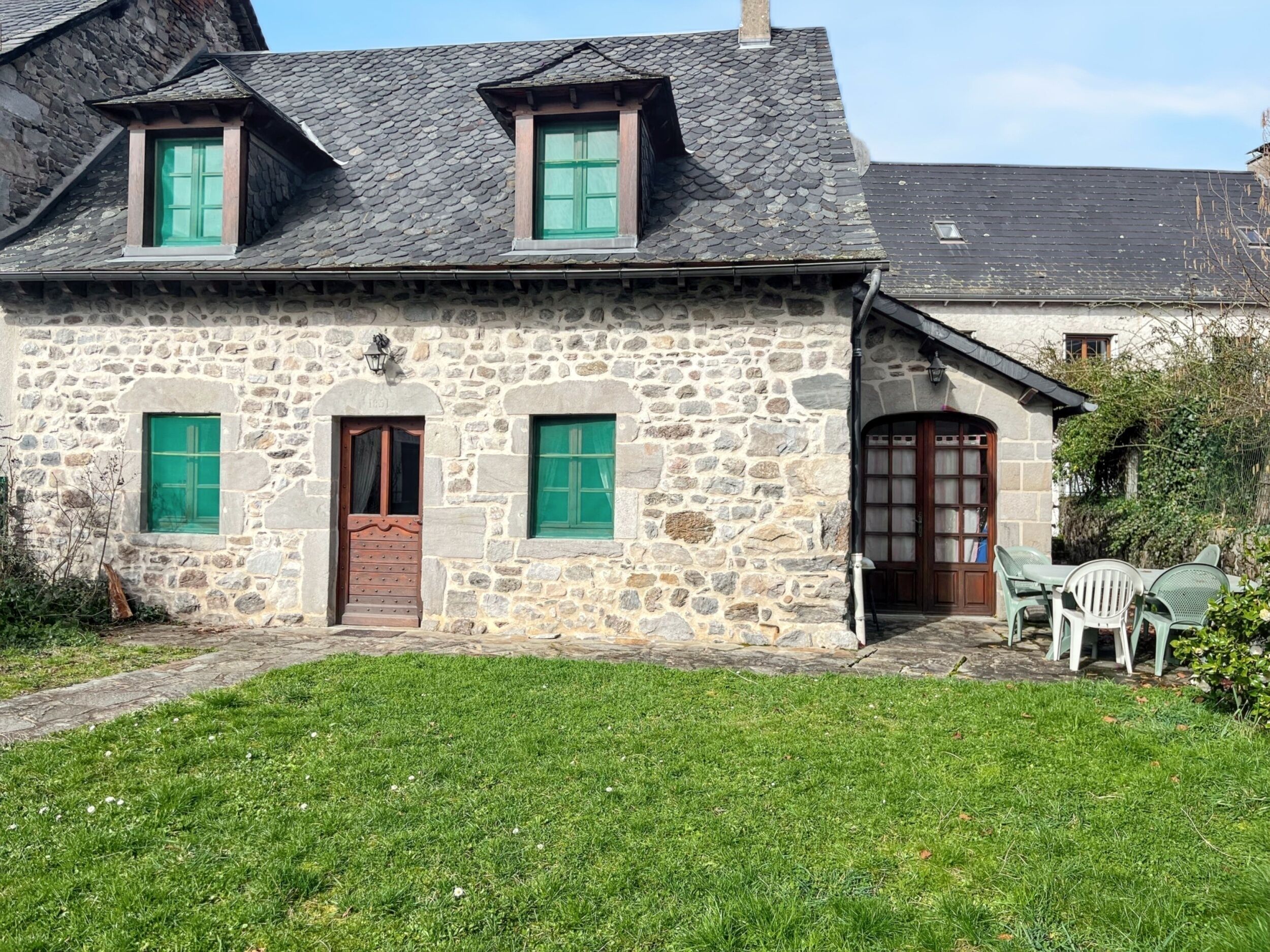 Una casa de piedra con puerta verde, dos ventanas con contraventanas verdes y un pequeño porche con mobiliario exterior.