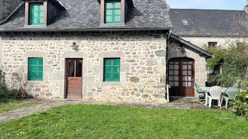 Una casa de piedra con puerta verde, dos ventanas con contraventanas verdes y un pequeño porche con mobiliario exterior.