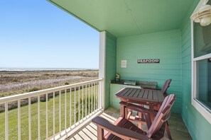 A balcony with a view of the beach, a sign indicating seafood availability, and a table and chairs set up for dining. - Knot 4 Sail -Beach Front- Ocean Views- Front Row - 2BD/2BA. Sleeps 6 (Galveston)