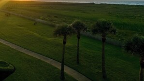 A grassy field with a path and palm trees, leading to a body of water with a sunset in the background. - Knot 4 Sail -Beach Front- Ocean Views- Front Row - 2BD/2BA. Sleeps 6 (Galveston)