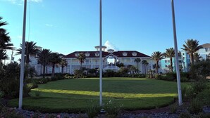 A flagpole with the American flag, Texas state flag, and a white flag. In the background, there is a large building with a red roof and multiple windows. - Knot 4 Sail -Beach Front- Ocean Views- Front Row - 2BD/2BA. Sleeps 6 (Galveston)