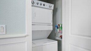 A laundry room with a stacked washer and dryer, a shelf with cleaning supplies, and a laundry control panel. - Knot 4 Sail -Beach Front- Ocean Views- Front Row - 2BD/2BA. Sleeps 6 (Galveston)