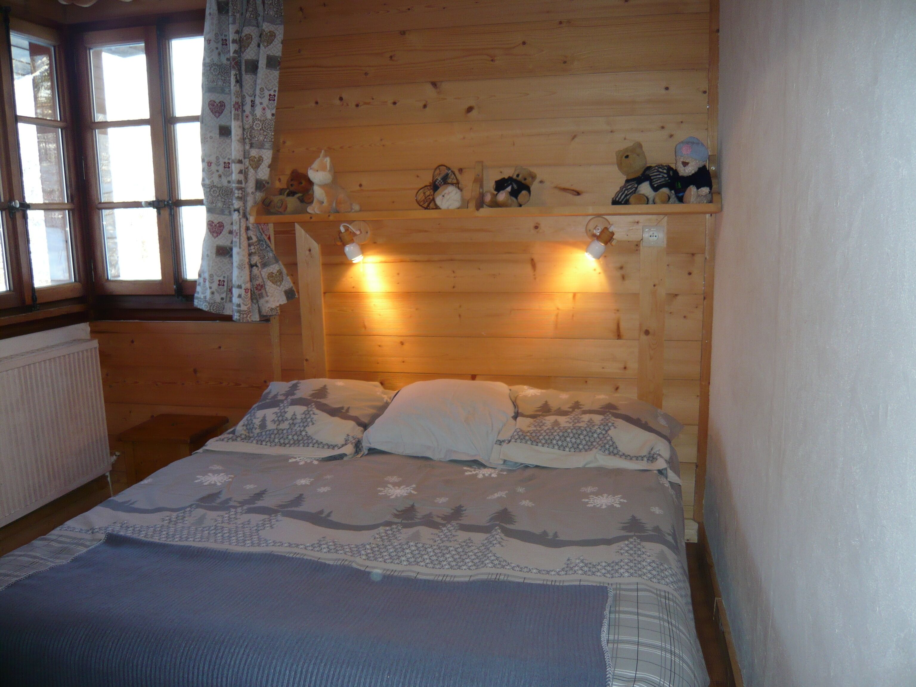 A wooden cabin bedroom with a bed, a shelf with stuffed animals, and a window with curtains.