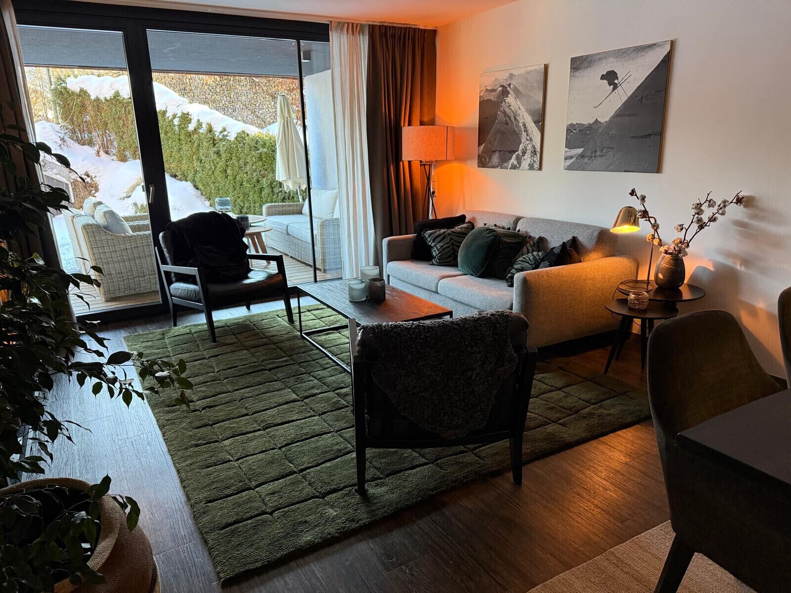 Ein modernes Wohnzimmer mit einer Couch, Sesseln, einem Couchtisch und einem Esstisch. Ein Schiebetürfenster führt in einen Außenbereich mit Blick auf schneebedeckte Bäume.