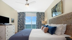A bedroom with a large bed, a ceiling fan, a television, and a balcony with curtains. - Lighthouse 608 - Great Location on the Gulf-Gulf Shores-Signature Properties (Gulf Shores)