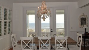 A dining room with a view of the ocean through a glass door, a chandelier, and white furniture.