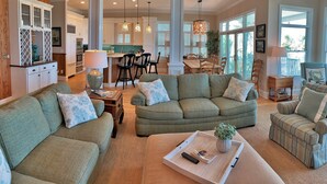 A spacious living room with green sofas, a dining area with a wooden table, and a kitchen in the background. - OCEANFRONT / Sleeps 14 / BISHOPS / POOL / ELEVATOR / SUMMER KITCHEN (Isle of Palms)