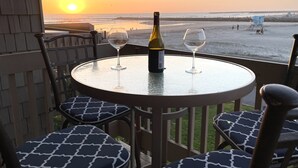 Een ronde tafel met twee wijnglazen en een fles, met uitzicht op een strand bij zonsondergang.