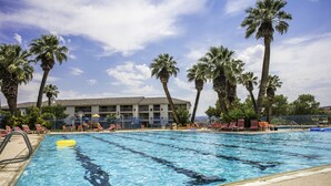 A swimming pool with clear blue water, surrounded by palm trees and a building with multiple windows in the background. - Spacious Family Condo  Ground Floor, Patio, Pools & Bikes & Fun All YEAR-ROUND  (St George)