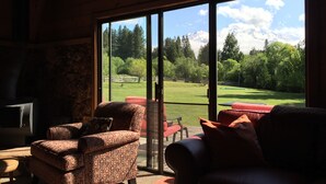 A room with a large sliding glass door leading to a view of a golf course and mountains. Inside, there are two patterned armchairs and a small table. - Amazing property & Shasta views, private lake, dark skies, close to everything (Mt. Shasta)