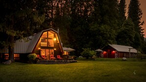 A large, A-frame wooden cabin with a porch and outdoor seating area, surrounded by a grassy field and trees. - Amazing property & Shasta views, private lake, dark skies, close to everything (Mt. Shasta)