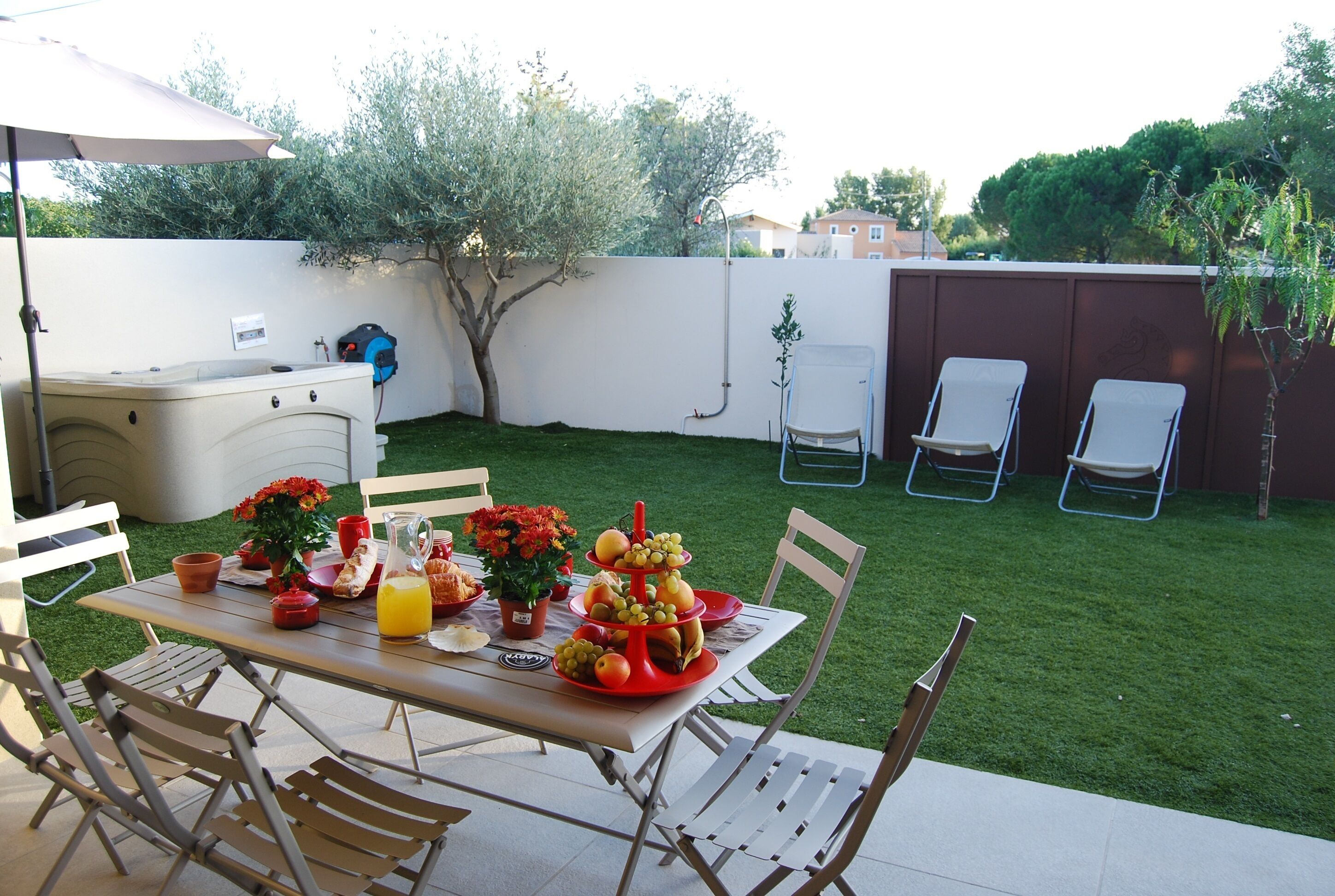 Un jardin aménagé avec une table à manger, des chaises disposées autour, et un petit espace potager avec un arbre et un tuyau d’arrosage.