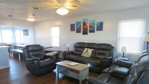A living room with a ceiling fan, a dining area with a table and chairs, and artwork on the wall. - Last minute special/Ocean Front/ Pool, Pool Table/pet friendly (Nags Head)