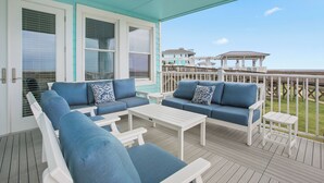 A covered outdoor seating area with blue sofas and white tables, overlooking a beach and a gazebo.