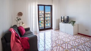 Un salon moderne avec un canapé, un téléviseur posé sur un meuble blanc, un sol à motifs et une vue sur les immeubles à travers une porte vitrée.