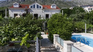 Une maison à deux étages, avec un toit recouvert de tuiles rouges, une piscine et une vue sur les montagnes en arrière-plan.