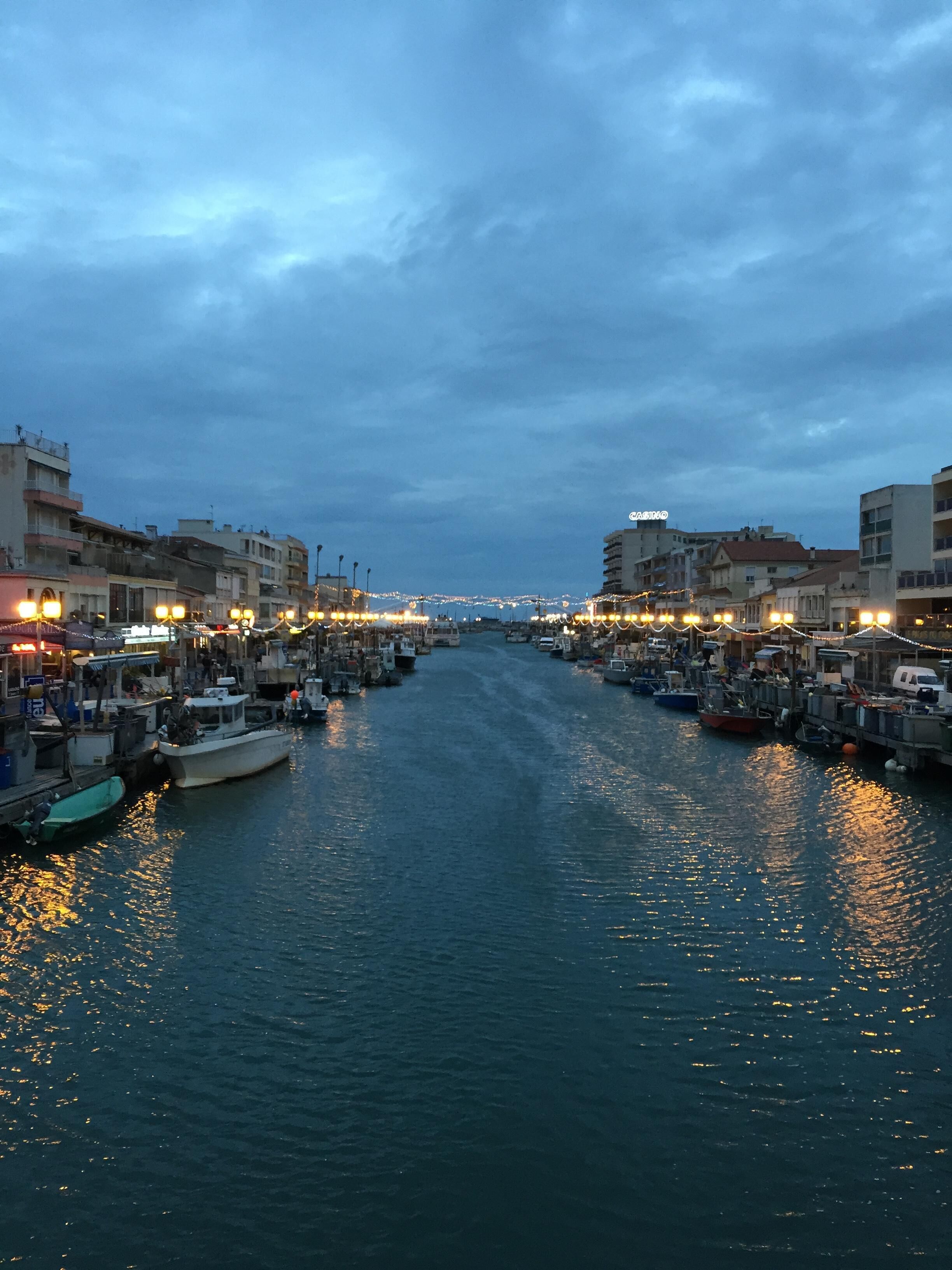 A narrow waterway lined with boats and buildings, reflecting lights on the water.