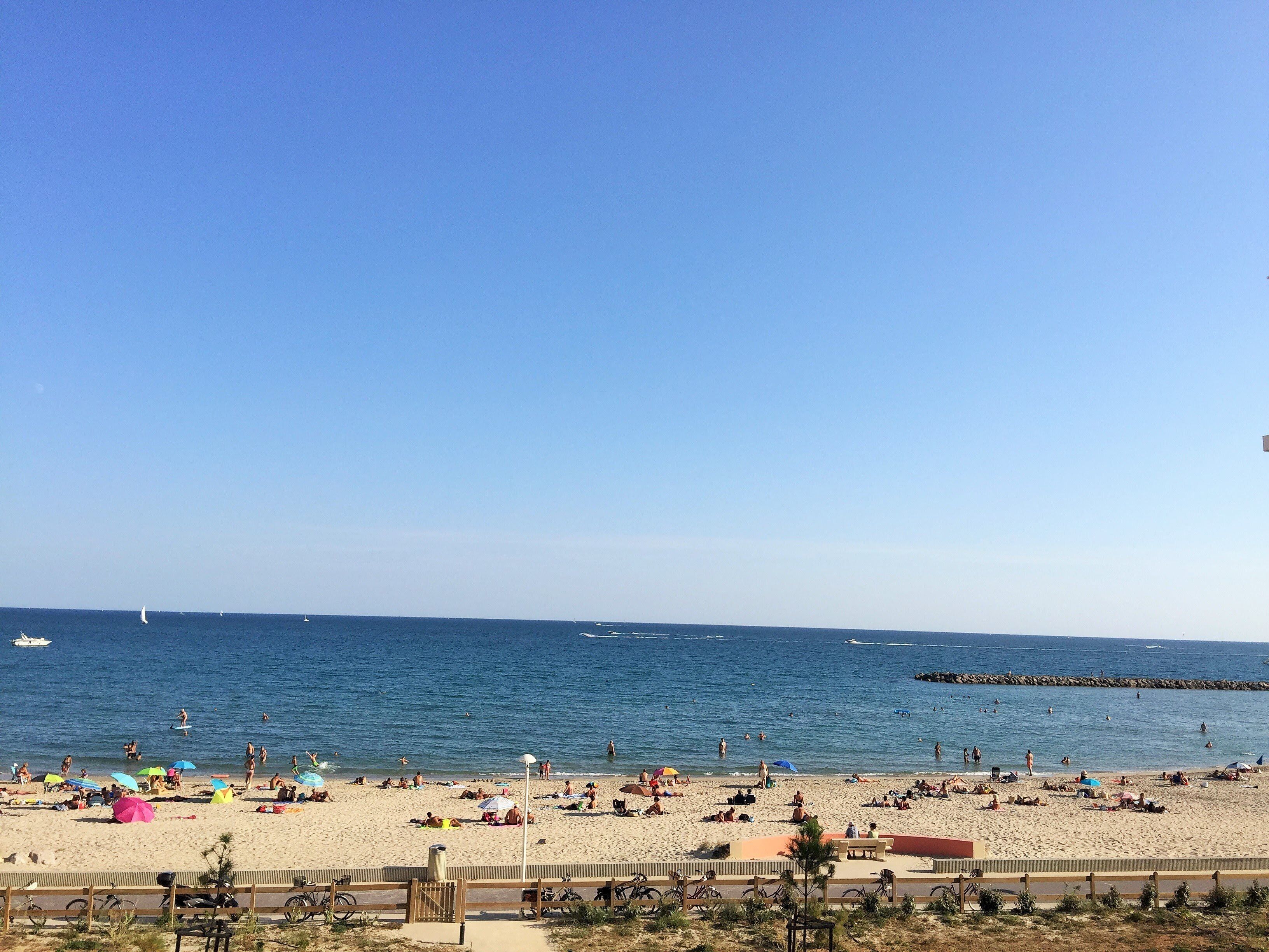 A beach with people sunbathing and swimming, a clear blue sky, and a rocky breakwater in the distance.