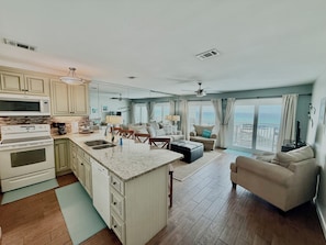 A modern kitchen with a central island, a dining area with a table and chairs, a living room with a sofa and a TV, and large windows with a view of the ocean.