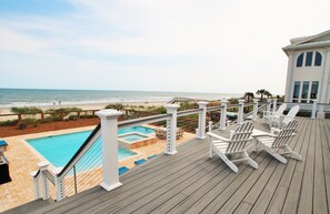 Terraza de madera con piscina, vista a la playa y sillones Adirondack blancos.