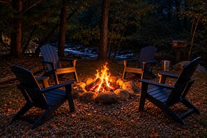 Lejrild med fire Adirondack-stole placeret omkring det, anlagt på et lag småsten med en flod og træer i baggrunden.