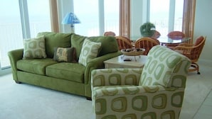 A living room with a green sofa, patterned armchair, wicker chairs, and a small table with a lamp.