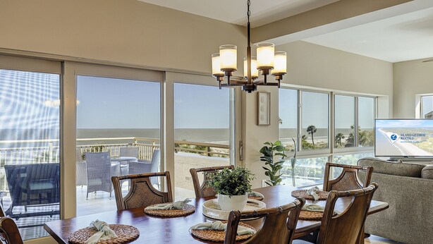 Uma sala de jantar com vista para a praia, um candelabro e uma televisão exibindo 'Island Getaway Rentals'.