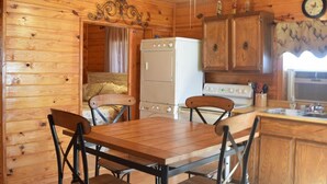 A wooden cabin interior with a dining table, chairs, a refrigerator, a stove, and wooden cabinets.