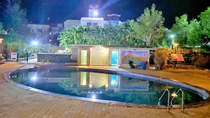 Un área de piscina nocturna con una piscina cristalina, un edificio pequeño con un mural y vegetación circundante.
