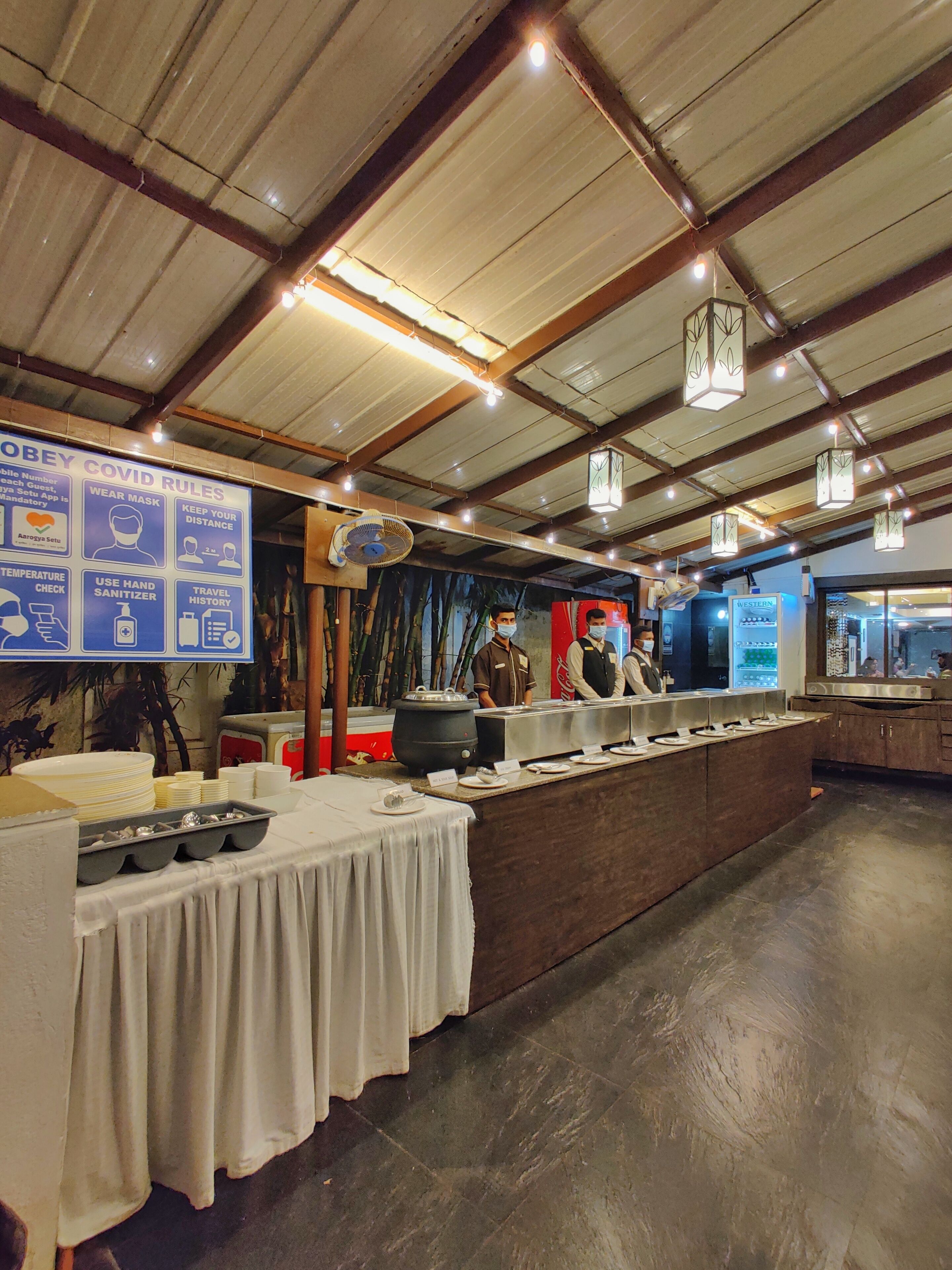 Un restaurante espacioso con techo alto e iluminación de estilo industrial. Un gran panel muestra las normas de seguridad por COVID-19.