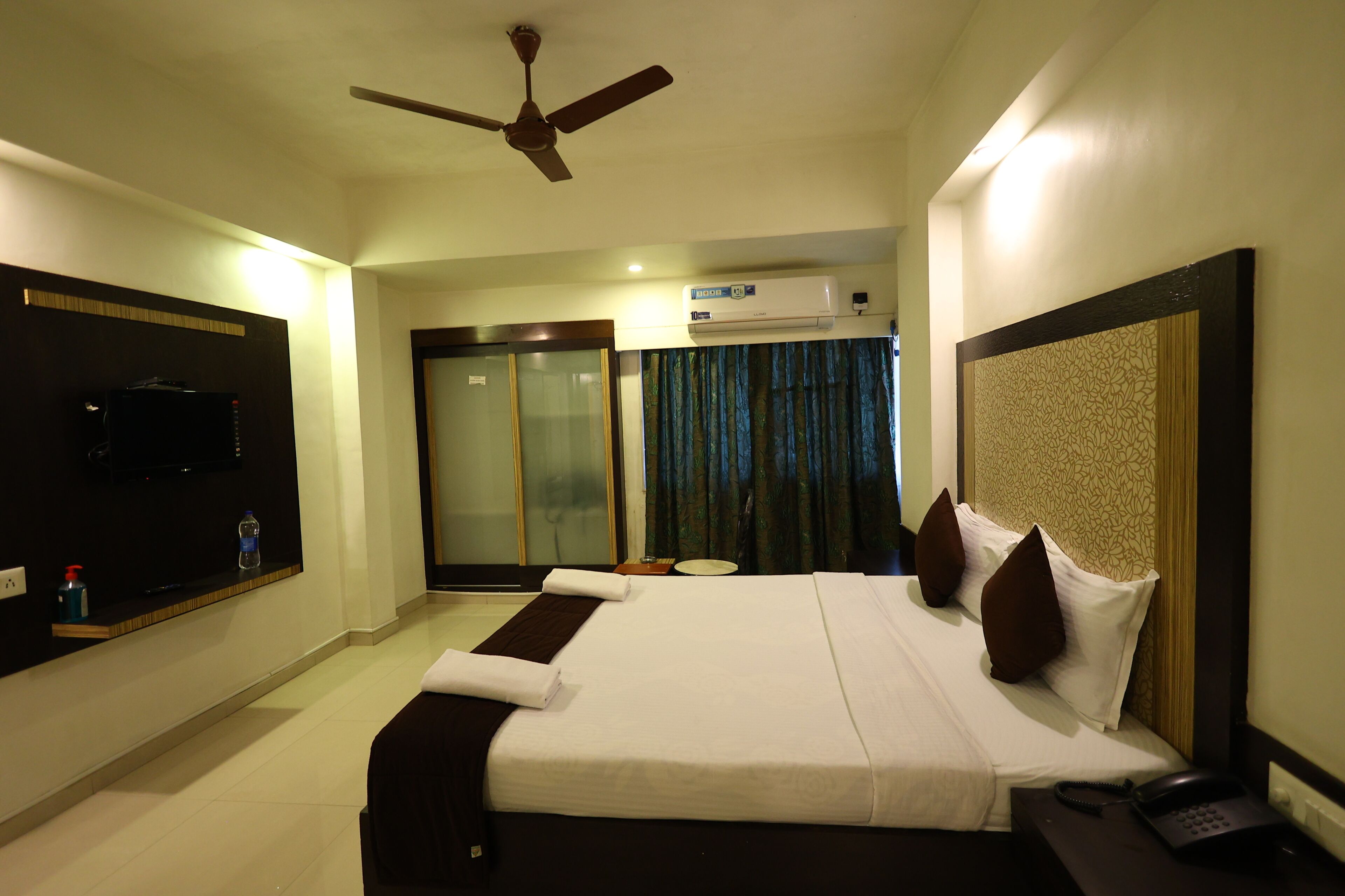 A hotel room with a bed, a ceiling fan, a TV, an air conditioning unit, and a bedside table with a phone.