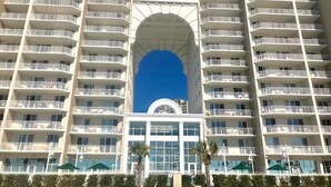 Un hôtel à plusieurs étages, doté d’une entrée centrale en forme d’arche, de balcons à chaque étage et, en arrière-plan, d’un ciel d’un bleu éclatant.