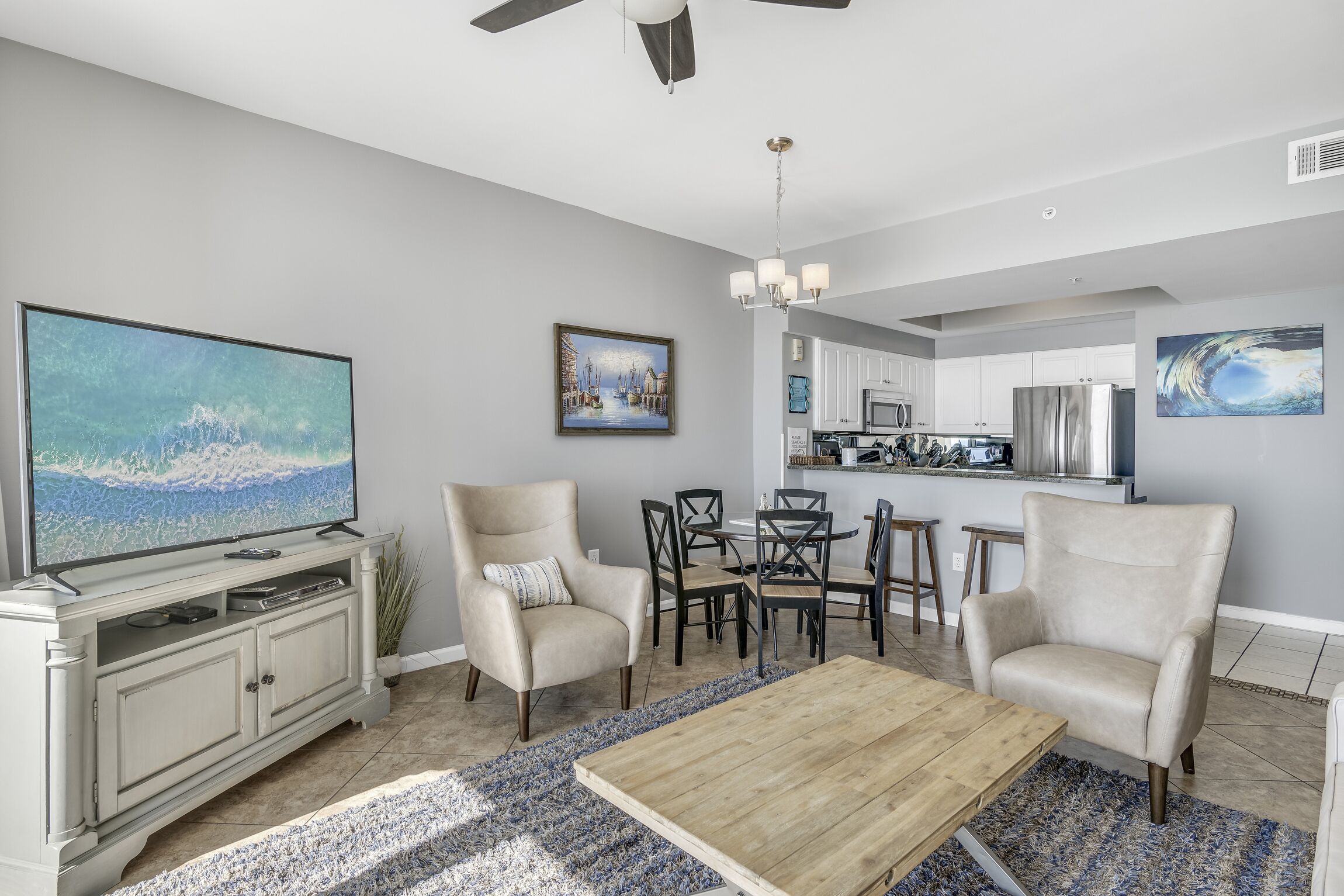 A modern living room with a flat-screen TV, beige armchairs, a wooden coffee table, and a dining area with a chandelier.