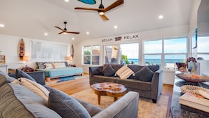 A spacious living room with a large gray sofa, a wooden coffee table, and a ceiling fan. A large window offers a view of the ocean. - Beachfront | Sunsets | Spa | Surf Spot (Haleiwa)