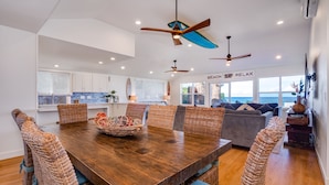A spacious dining area with a wooden table and wicker chairs, a kitchen in the background, and a view of the ocean through large windows. - Beachfront | Sunsets | Spa | Surf Spot (Haleiwa)