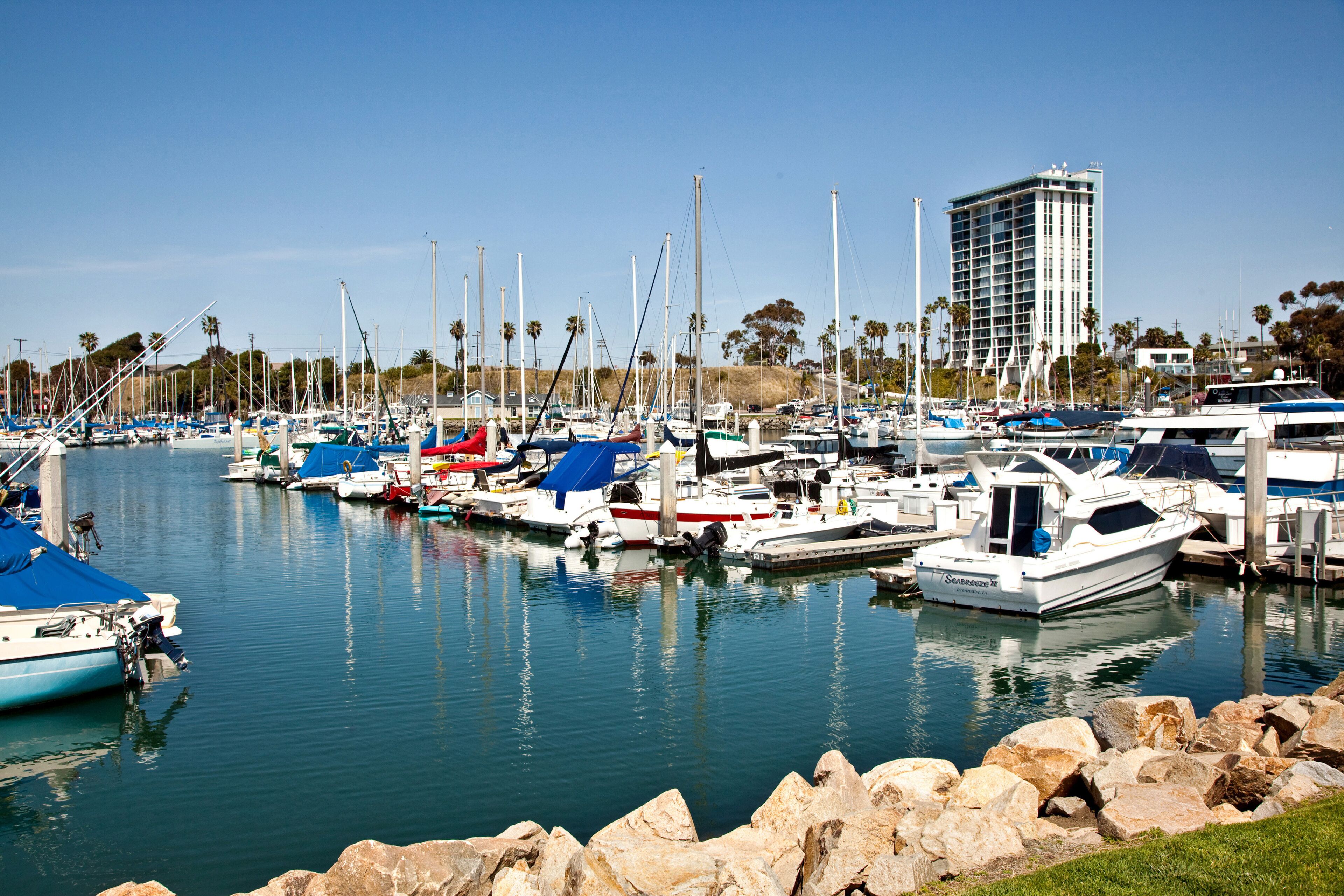 A marina with various boats docked, a large building in the background, and a rocky shoreline.