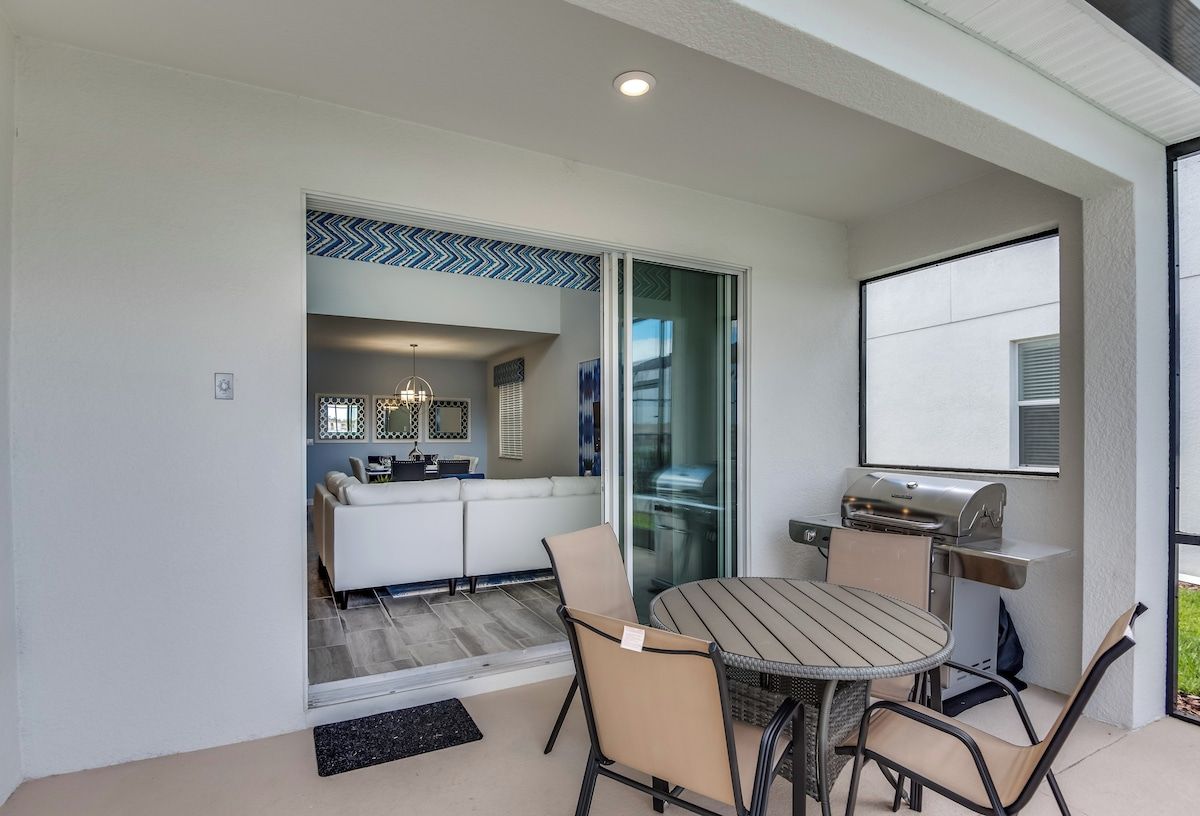 A patio area with a round table and chairs, a grill, and a sliding glass door leading to a living room.
