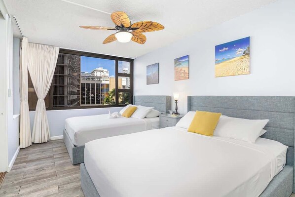 A hotel room with two beds, a ceiling fan, a large window with a view of buildings, and artwork on the wall. - Elegant 1BR condo w/ pool & free parking (Honolulu)