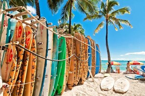 A row of surfboards leaning against a structure on a beach with palm trees and umbrellas in the background. - Elegant 1BR condo w/ pool & free parking (Honolulu)