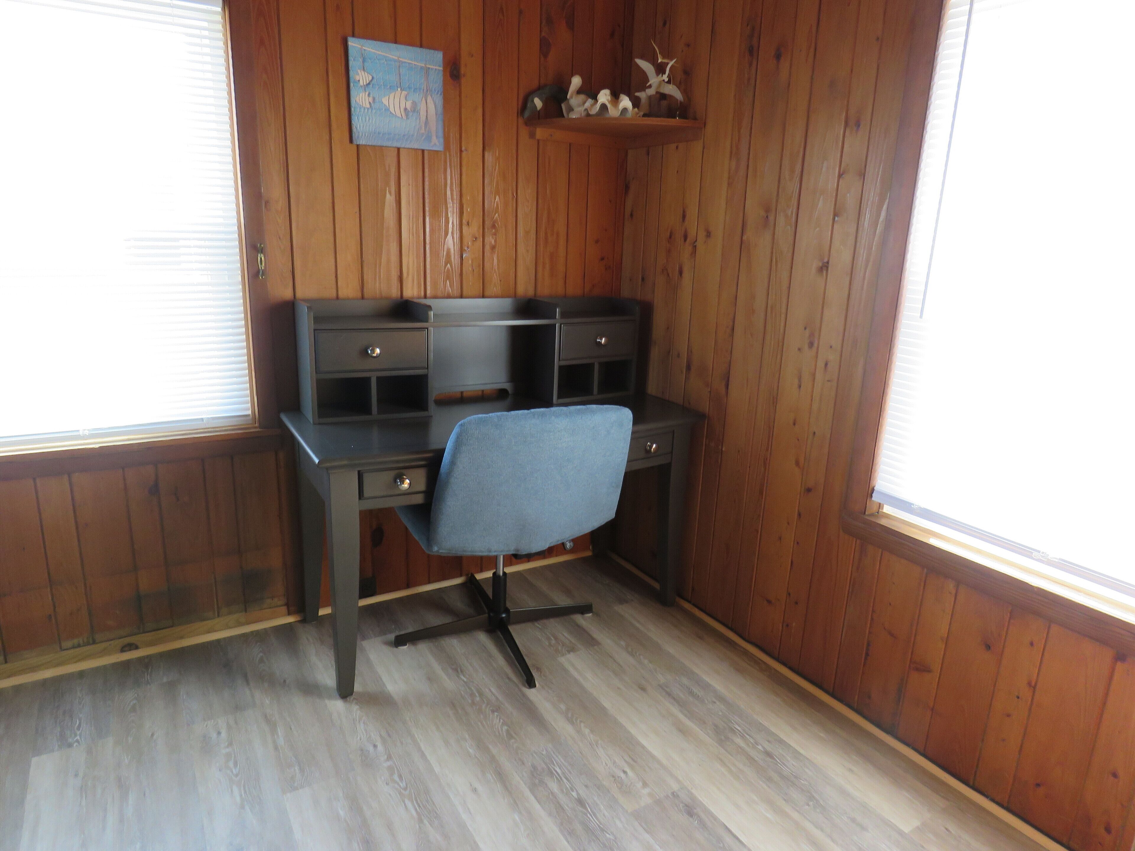 Une pièce d’angle avec un mur lambrissé en bois, un bureau avec des tiroirs, une chaise et une petite étagère ornée d’objets décoratifs.