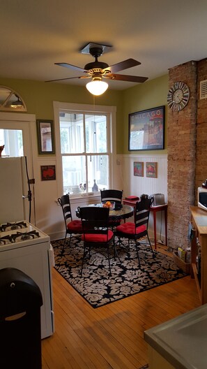 A kitchen with a dining table, chairs, a ceiling fan, a refrigerator, and a brick wall.