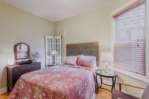 A bedroom with a bed, dresser, bedside table, lamp, and window with blinds.