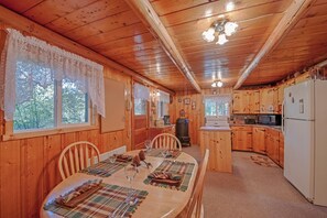 A wooden cabin with a dining area, kitchen, and a large window with lace curtains.