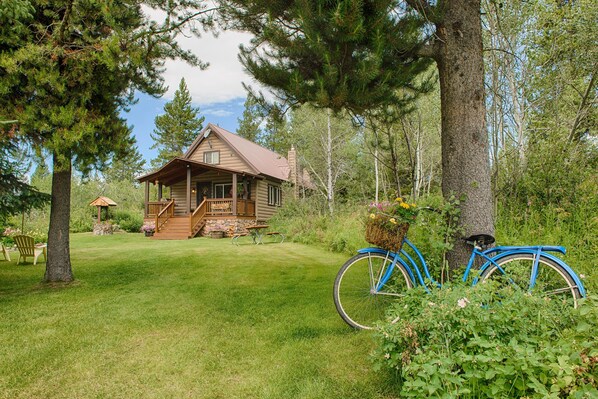 A log cabin with a porch, surrounded by trees and a grassy yard.
