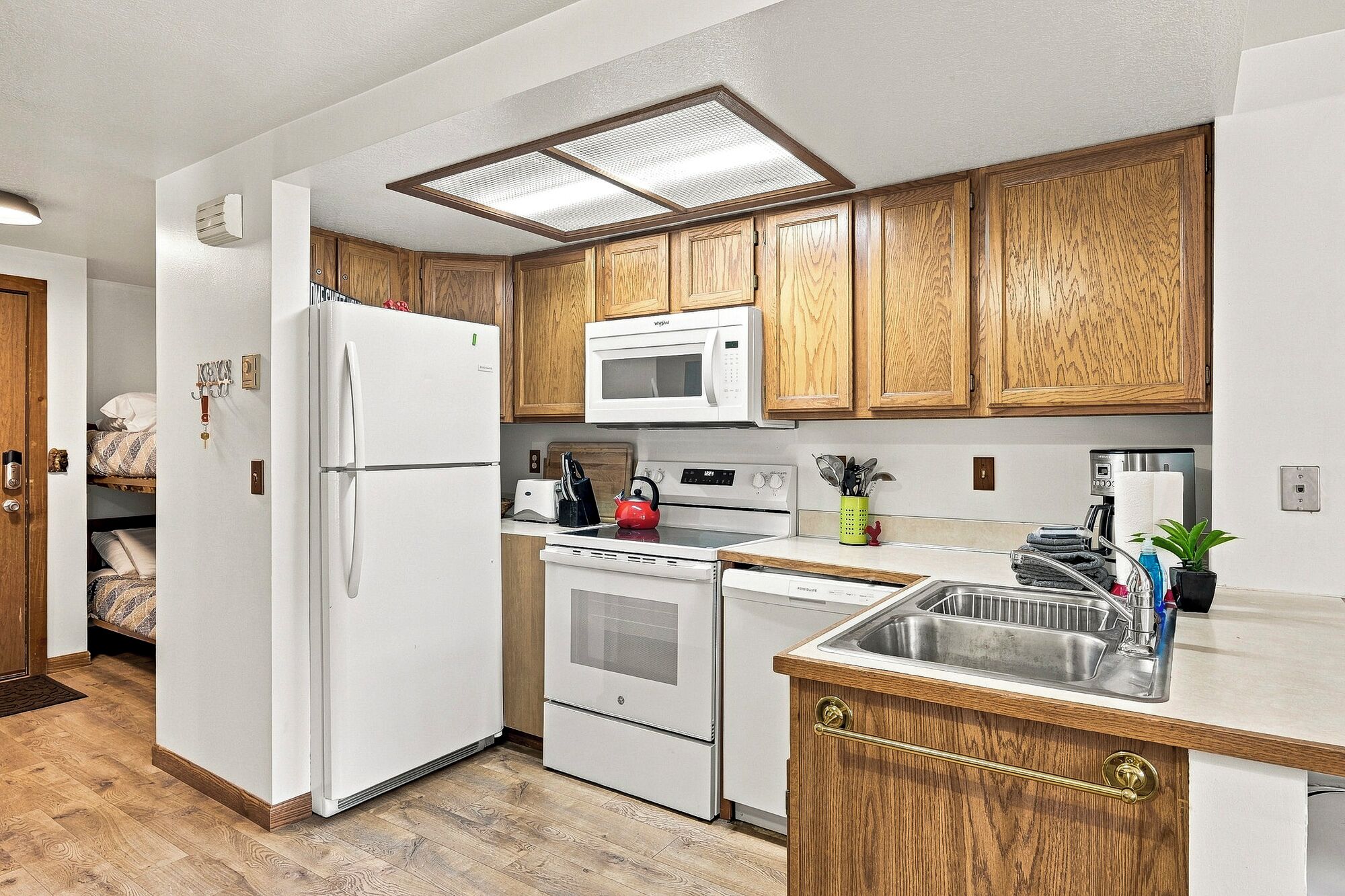 A compact kitchen with wooden cabinets, a white refrigerator, a microwave, a kettle, and a stainless steel sink.