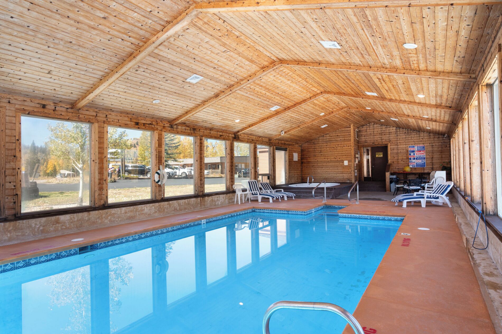 An indoor swimming pool with clear windows, lounge chairs, and a wooden ceiling.