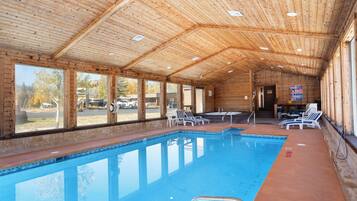 An indoor swimming pool with clear windows, lounge chairs, and a wooden ceiling.