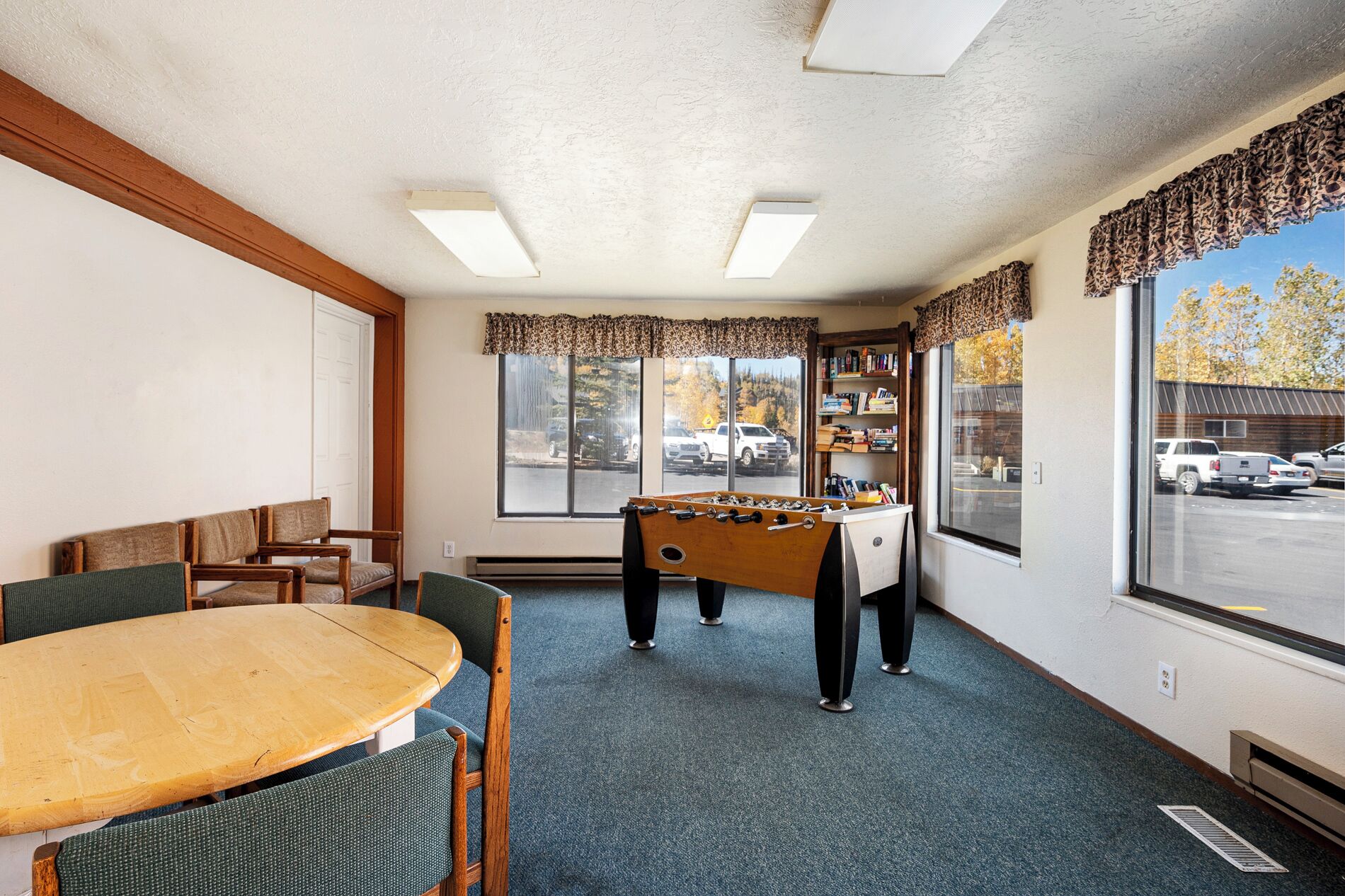 A room with a foosball table, a dining table with chairs, and large windows with a view of the outside.